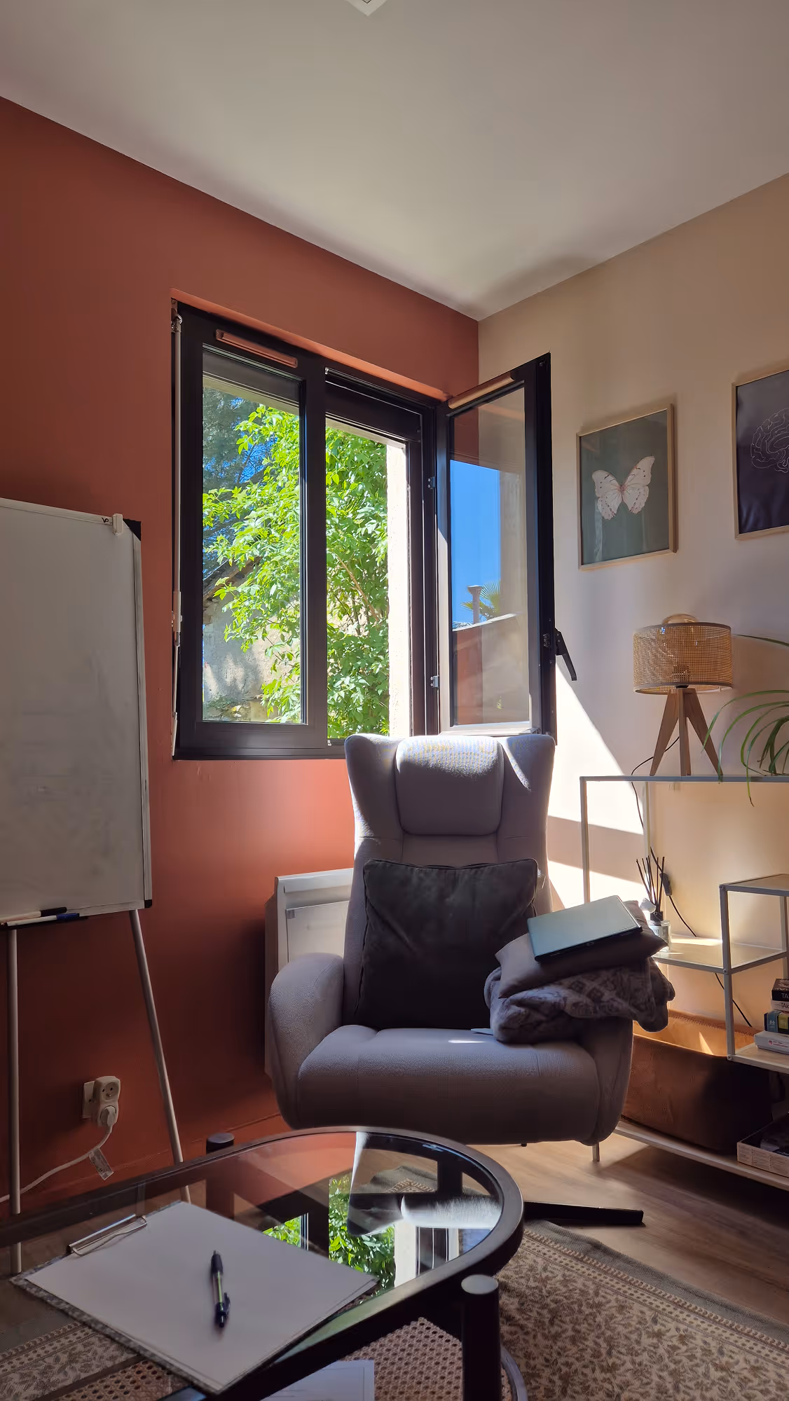 Cabinet de Margot Daubas, psychologue TCC à la Possonnière, près d'Angers. Une pièce confortable avec un fauteuil et un canapé, une table en verre, une fenêtre ouverte, un tableau blanc, une lampe et des tableaux aux murs.