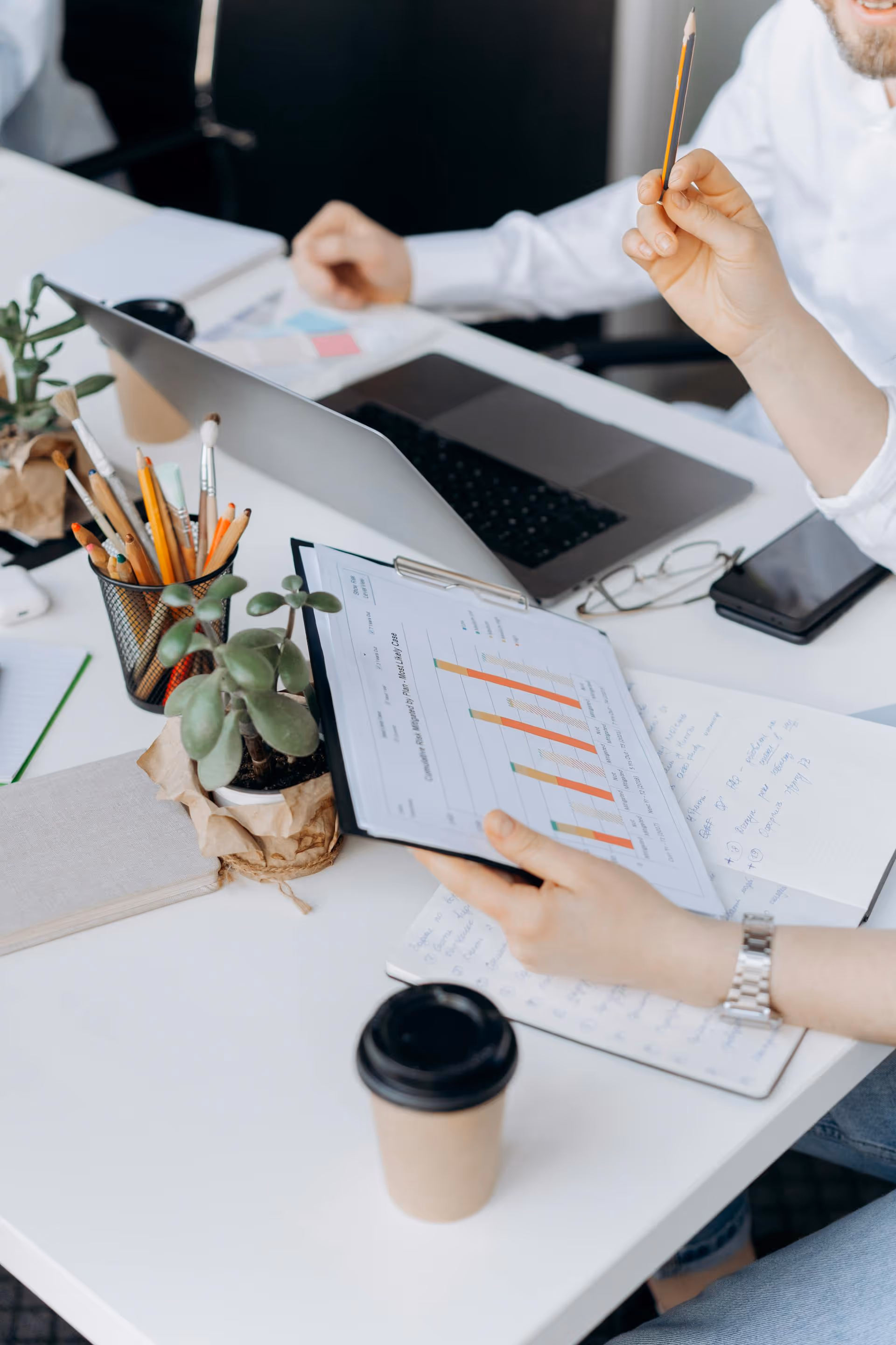 Une personne tenant un bloc-notes avec un graphique à barres, travaillant à un bureau avec un ordinateur portable, des plantes et une tasse de café.
